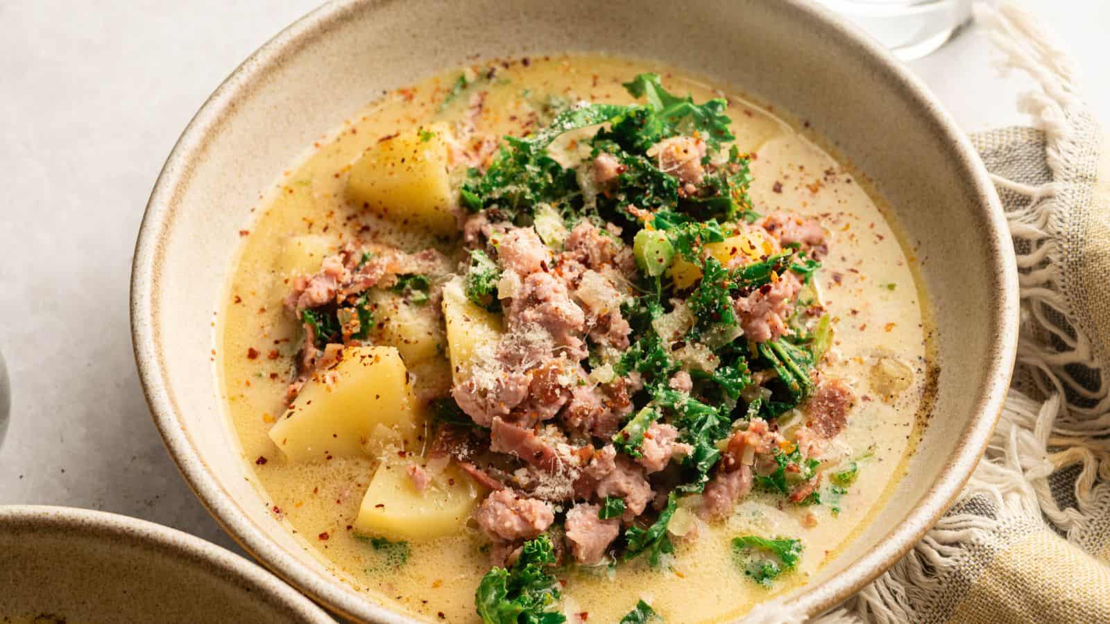 A bowl of creamy soup containing potatoes, crumbled sausage, kale, and sprinkled with grated cheese and red pepper flakes.