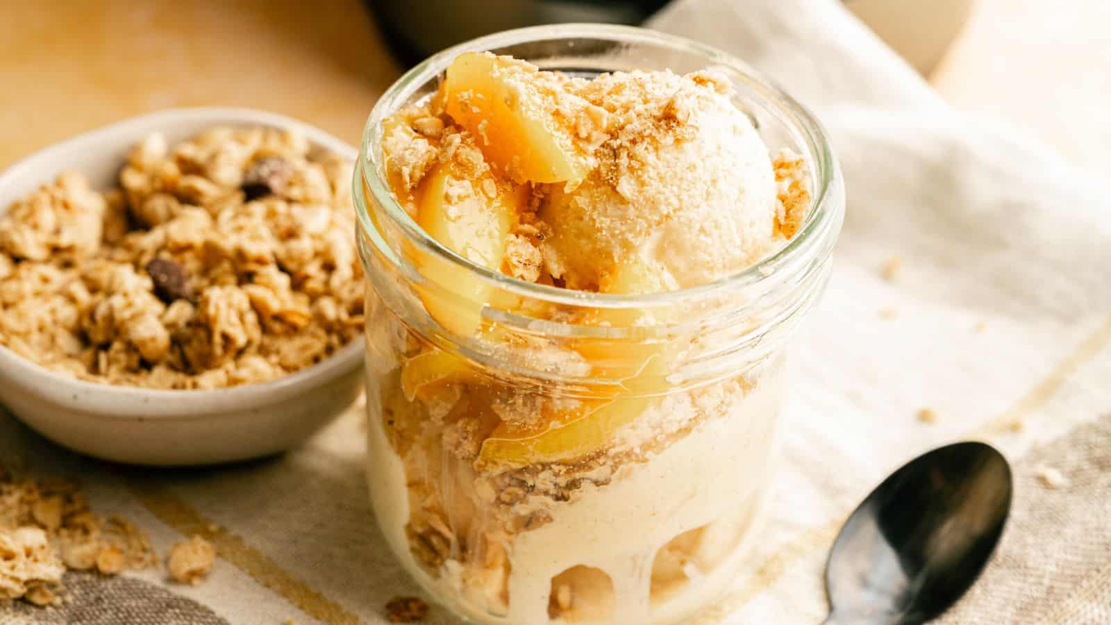 A glass jar filled with vanilla ice cream, peach slices, and granola sits on a cloth next to a bowl of granola and a spoon.