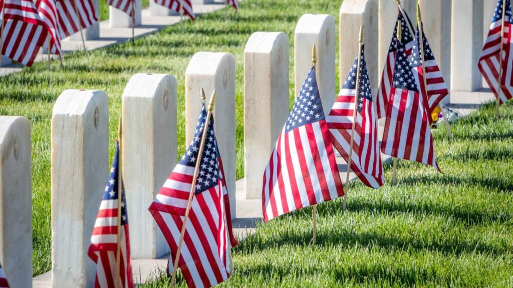 Rows of white gravestones with American flags placed in front, set in a cemetery with green grass, honor those remembered on Memorial Day.