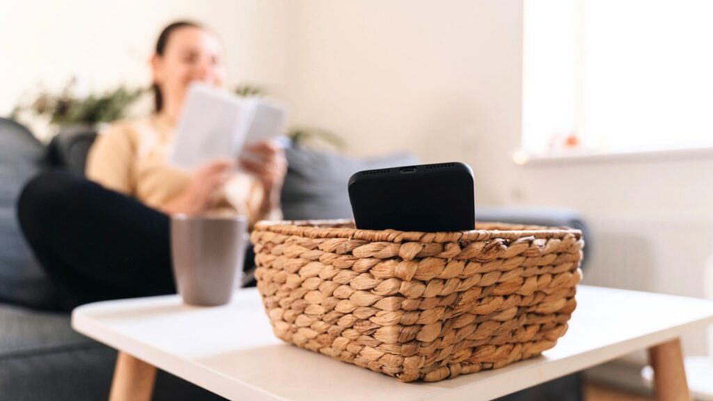 A smartphone in a wicker basket on a white table, with a person reading papers and holding a mug in the background on a couch&mdash;capturing a moment of calm away from over-optimization.