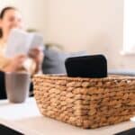 A smartphone in a wicker basket on a white table, with a person reading papers and holding a mug in the background on a couch—capturing a moment of calm away from over-optimization.