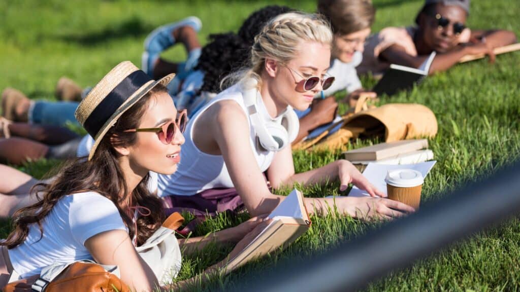A group of young adults lie on the grass, reading books and wearing sunglasses, with backpacks and a coffee cup nearby.