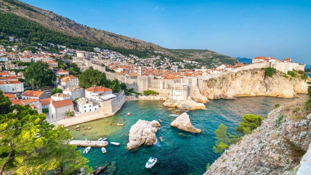 Aerial view of Dubrovnik&rsquo;s old town, one of the most stunning European cities, with stone city walls, red-roofed buildings, rocky coastline, and boats in the blue-green Adriatic Sea.