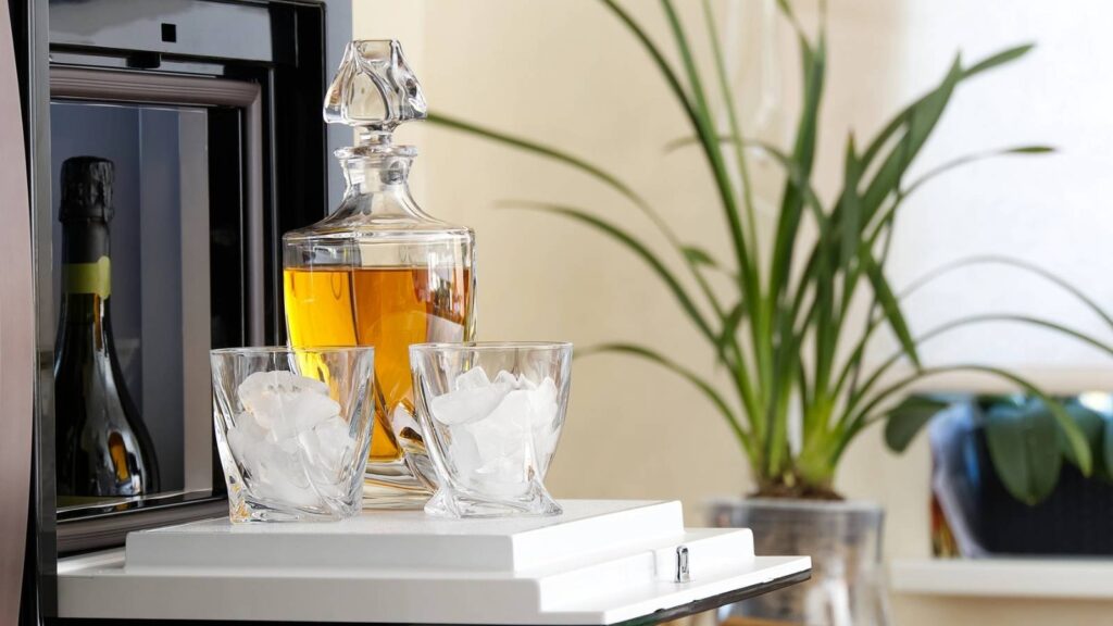 A decanter filled with amber liquid and two glasses with ice sit on a white tray beside a refrigerator, enhancing stylish bar areas with a wine bottle and a potted plant in the background.