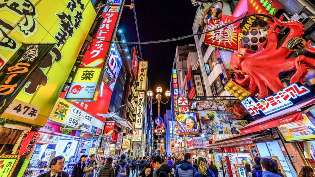 A busy street in Osaka, Japan at night, filled with bright neon signs, colorful advertisements, and crowds of people walking.
