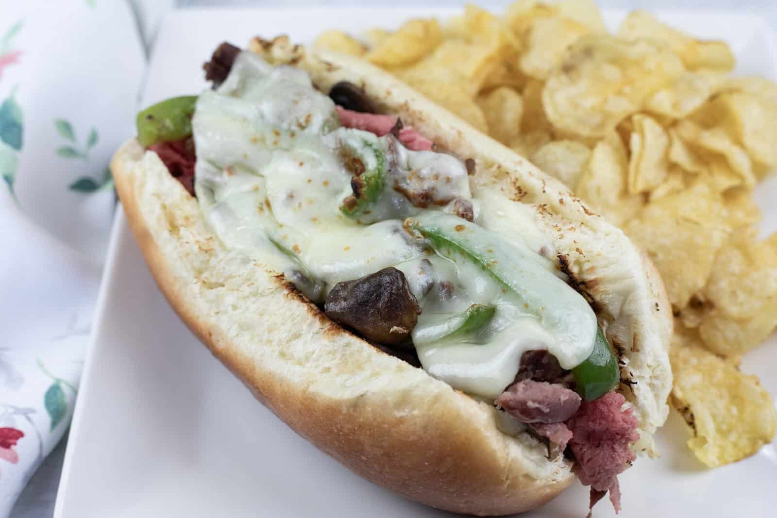 A Philly cheesesteak sandwich with melted cheese, green peppers, and onions in a hoagie roll, served with potato chips on a white plate.
