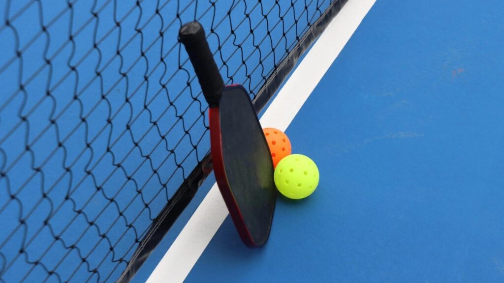 A pickleball paddle rests against a net on a blue court beside two perforated balls, one orange and one yellow, with a floating pickleball nearby.