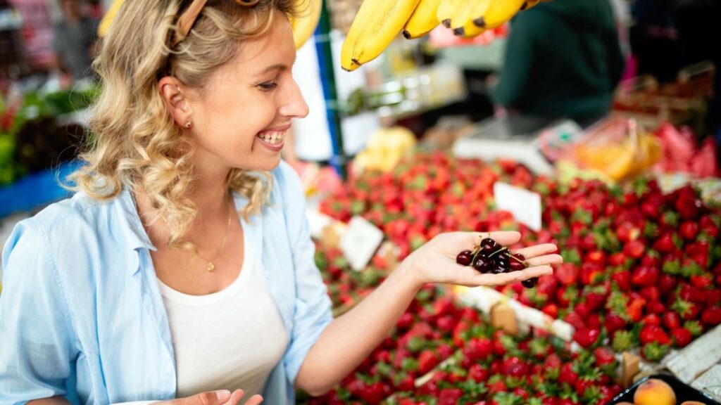 A woman holds dark cherries in her hand and smiles while standing in front of a vibrant farmers market fruit stall with strawberries and bananas.