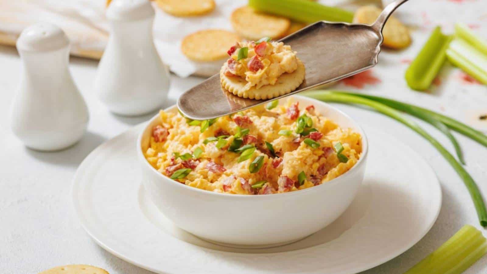A bowl of pimento cheese dip garnished with green onions, with a cracker topped with the dip being served. Crackers, celery sticks, and salt and pepper shakers are in the background.