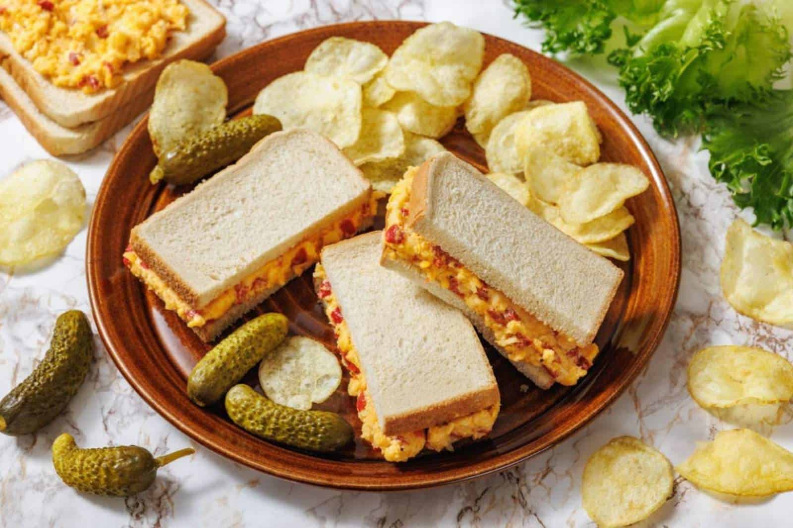 A plate with three pimento cheese sandwiches, potato chips, and pickles, with lettuce and more chips in the background.