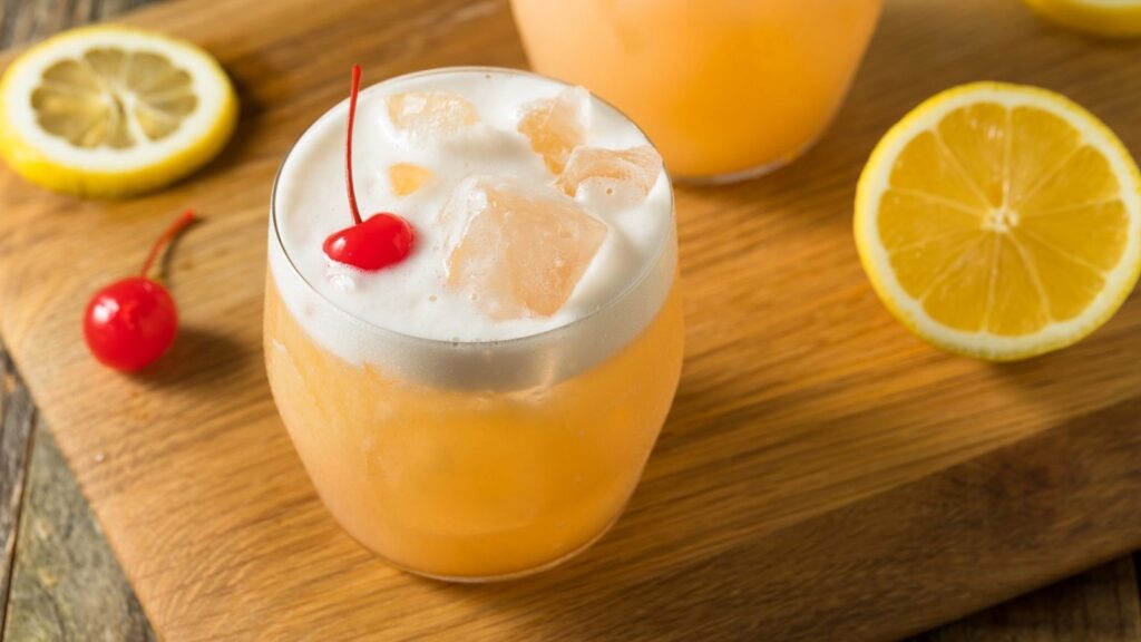 A glass of orange-colored cocktail with ice, a layer of cold foam on top, and a cherry garnish sits on a wooden board next to lemon slices.