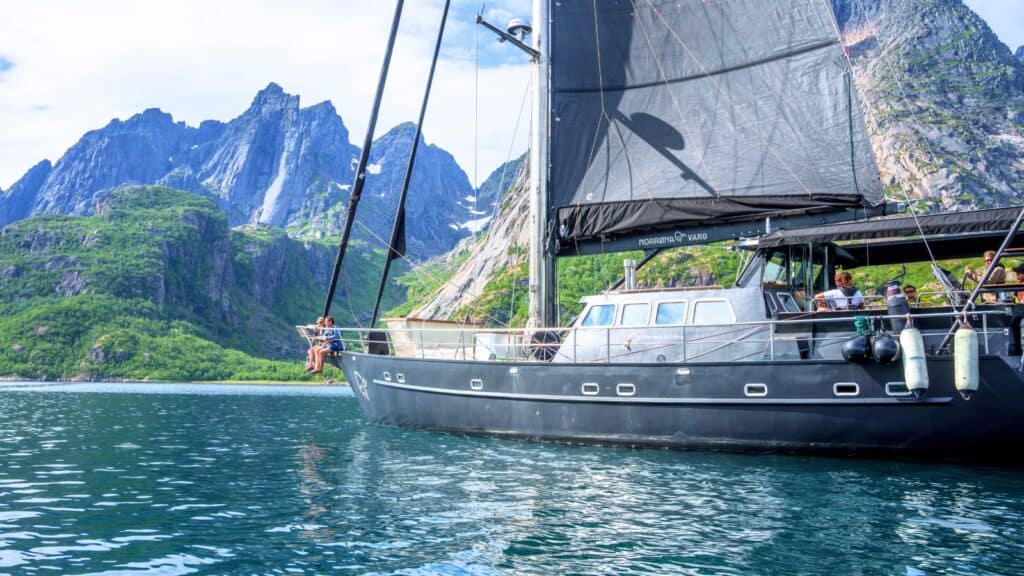 A black sailboat with people on board enjoys a luxury cruise on calm water near green mountains under a partly cloudy sky.