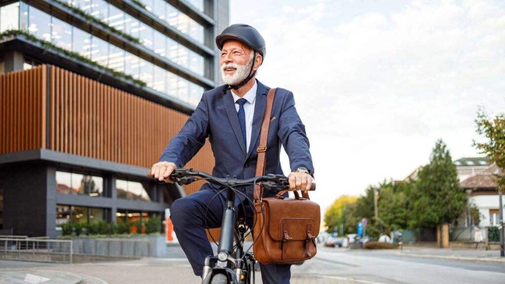 An older man in a suit and helmet rides a bicycle near modern office buildings, carrying a brown shoulder bag.