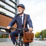 An older man in a suit and helmet rides a bicycle near modern office buildings, carrying a brown shoulder bag.