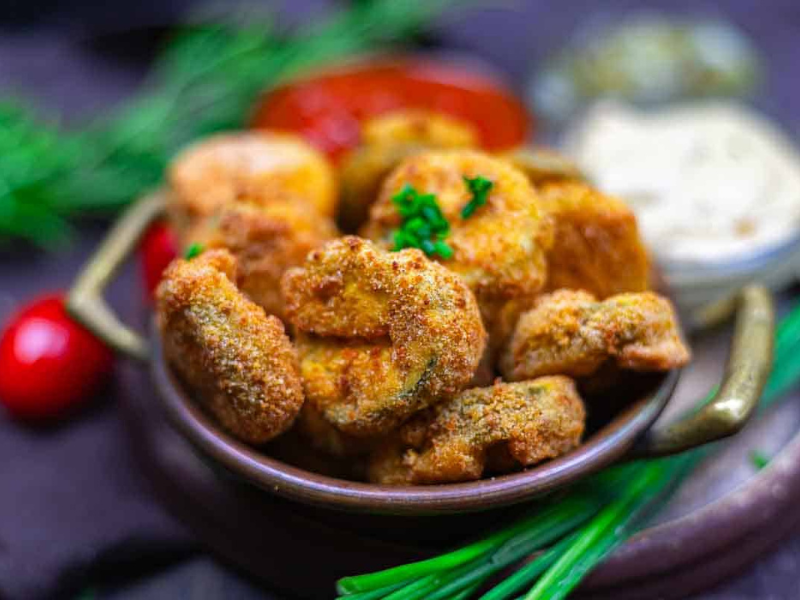 A bowl of breaded and fried pickle pieces garnished with parsley, with dipping sauces and herbs in the background.