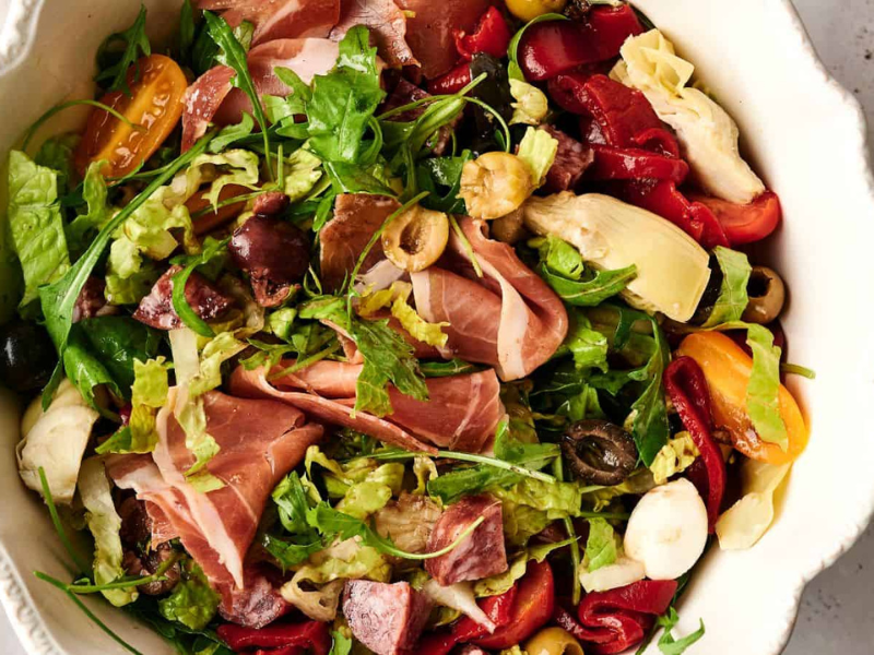 A bowl of salad with mixed greens, prosciutto, cherry tomatoes, olives, artichokes, and roasted red peppers.