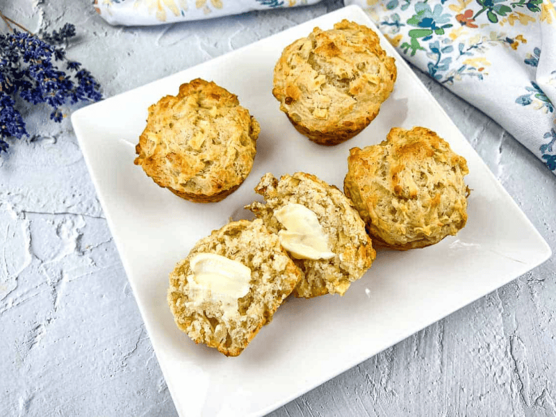 Apple cheddar muffins on a white plate, with one muffin cut open and spread with butter, showing the moist, cheesy interior.
