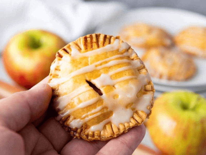 A hand holds a small round hand pie with icing drizzle; apples and more hand pies are visible in the background.