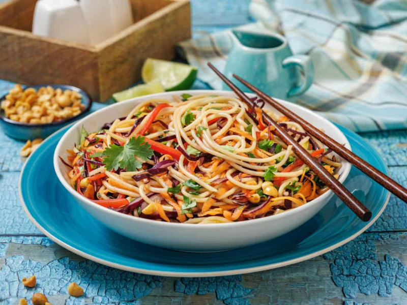 A bowl of colorful noodle salad with vegetables, topped with cilantro, placed on a blue plate with chopsticks, with peanuts and lime on the side.
