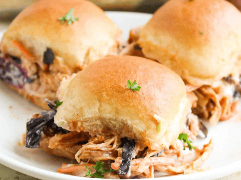 Three pulled pork sliders with coleslaw on a white plate, topped with a sprig of parsley.
