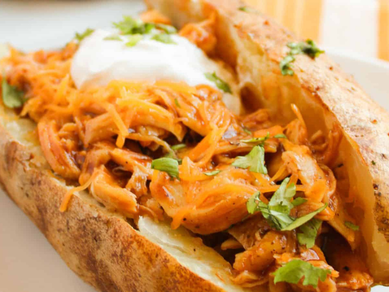 A baked potato topped with shredded barbecue chicken, sour cream, and chopped herbs on a white plate.