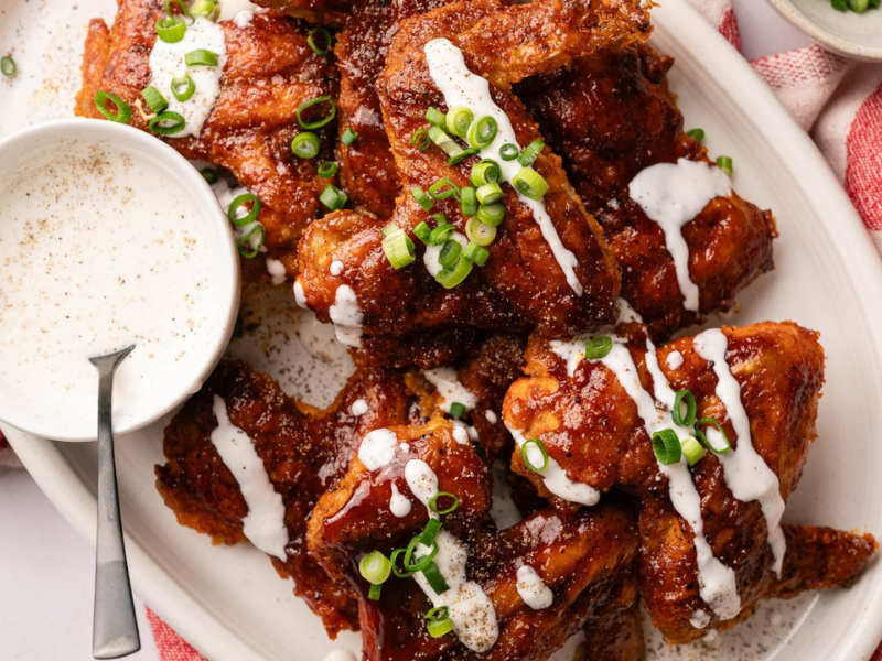 A platter of glazed chicken wings topped with chopped green onions and drizzled with white sauce, served with a small bowl of dipping sauce and a spoon.