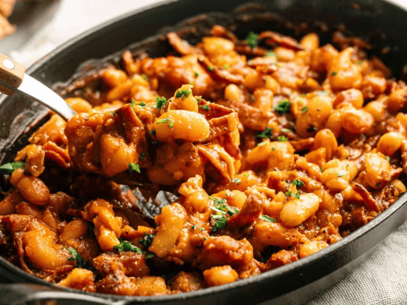 A black oval dish filled with baked beans in a rich sauce, mixed with pieces of bacon and garnished with chopped parsley.