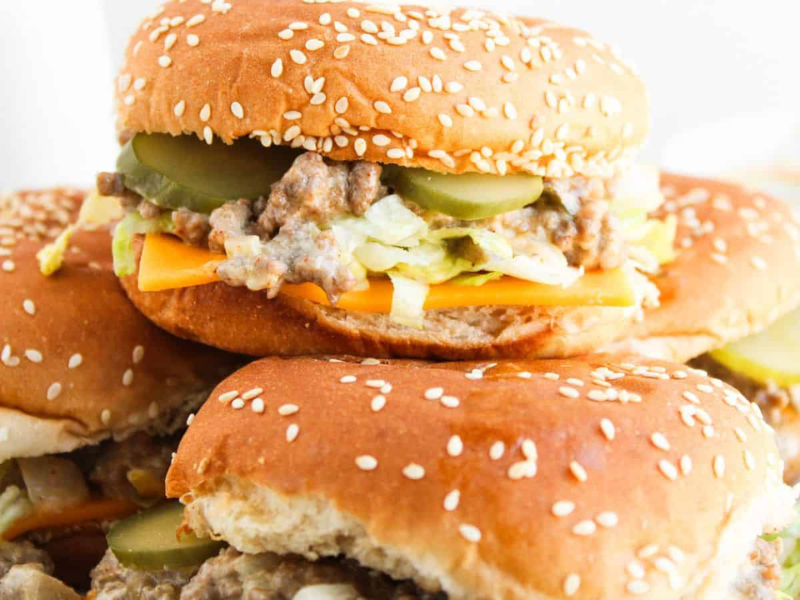 Two sesame seed burgers with cheese, shredded lettuce, ground meat, and pickles, stacked on a plate.