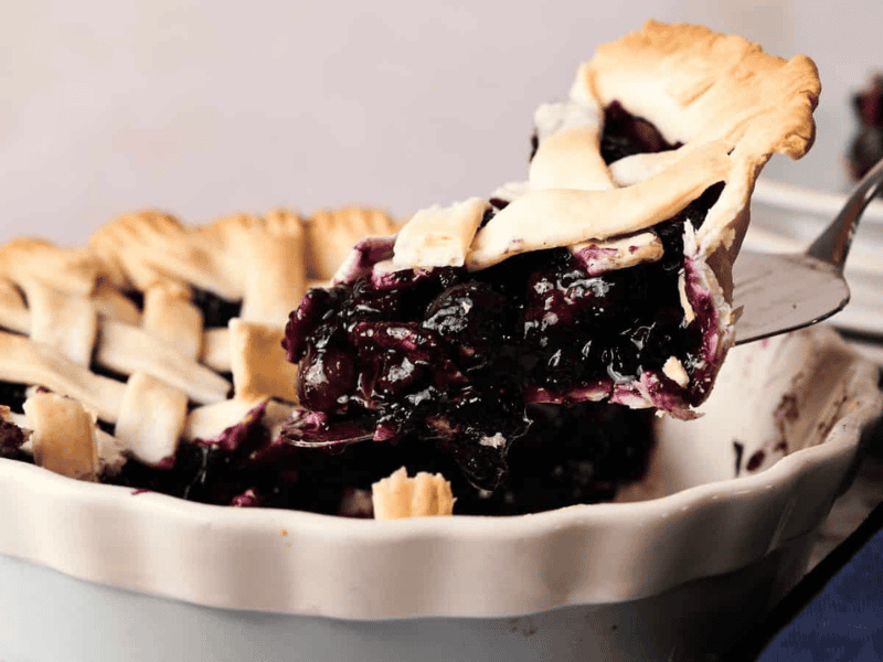 A slice of blueberry pie with a lattice crust is being lifted from a white ceramic pie dish.