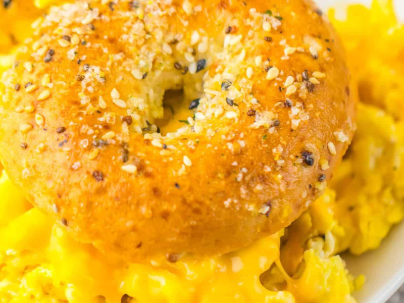 A sesame seed bagel sits on top of a serving of scrambled eggs with melted cheese on a white plate.
