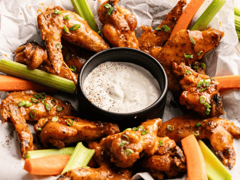 A tray of sauced chicken wings garnished with chopped green onions, served with celery and carrot sticks, and a cup of creamy dipping sauce in the center.