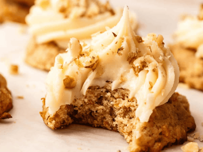 A frosted cookie topped with chopped nuts has a bite taken out of it; other similar cookies are visible in the background.