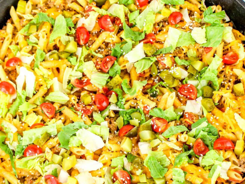 A skillet filled with penne pasta topped with shredded cheese, ground beef, diced tomatoes, lettuce, and pickles&mdash;a delicious take on cheeseburger pasta.