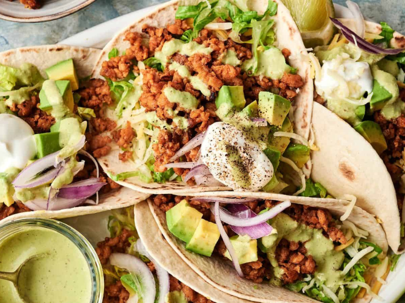 Three soft tacos filled with seasoned ground meat, shredded lettuce, diced avocado, red onion, shredded cheese, and topped with sour cream and green sauce sit on a plate.