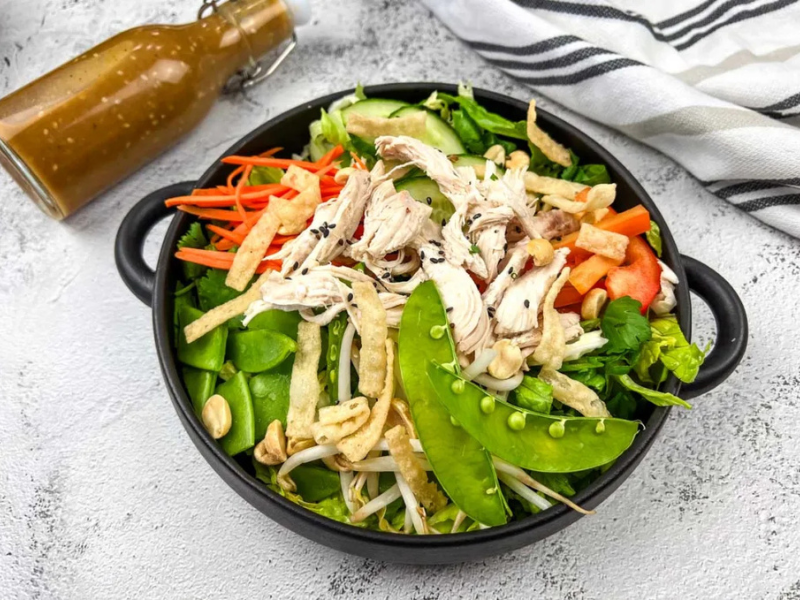 A bowl of Chinese chicken salad with shredded chicken, snap peas, carrots, and peppers, accompanied by a bottle of dressing on the side.