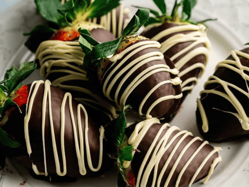 Six strawberries dipped in dark chocolate and drizzled with white chocolate are arranged on a white plate.