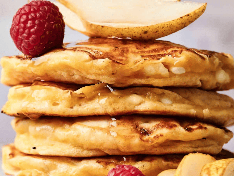 A stack of four fluffy pancakes topped with fresh raspberries, pear slices, and a drizzle of syrup.