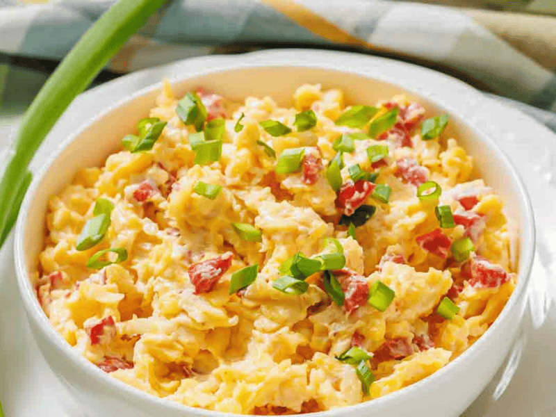 A bowl of pimento cheese spread with diced red bell peppers and topped with chopped green onions, set on a white plate.