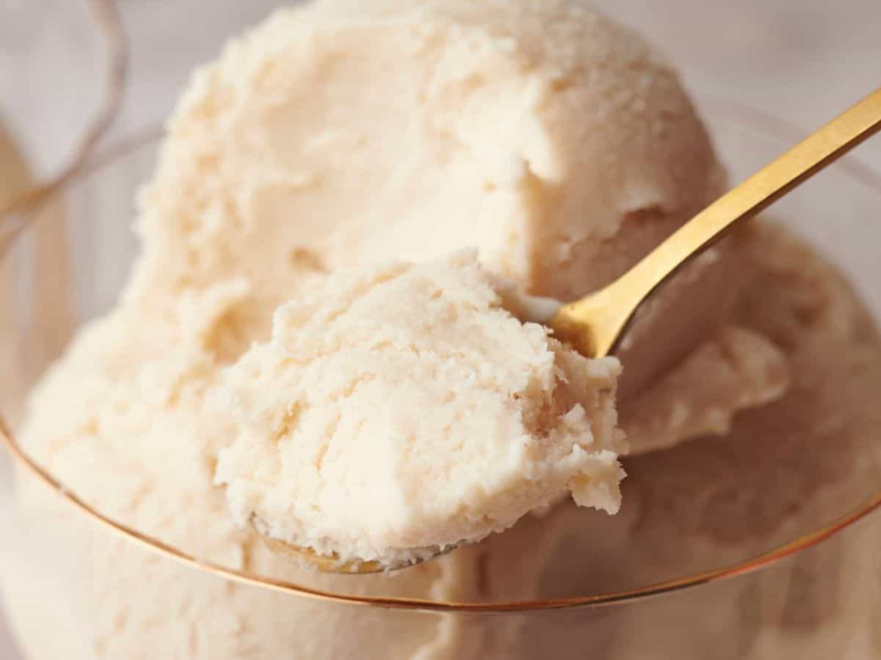 A gold spoon scoops a portion of creamy, pale vanilla ice cream from a glass bowl.