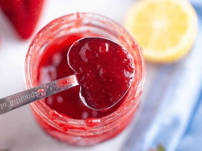 A heart-shaped measuring spoon holds a scoop of vibrant red strawberry puree over a glass jar filled with more puree, surrounded by fresh strawberries and a lemon half.