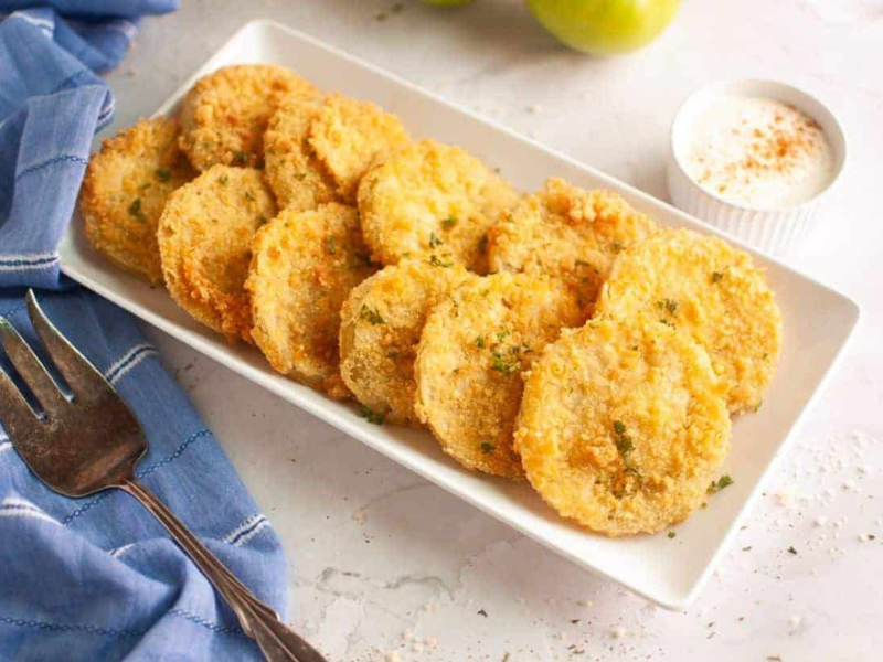 A rectangular white plate with fried green tomato slices garnished with herbs, a small cup of dipping sauce, a fork, and a blue napkin on the side.