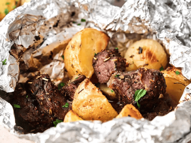 Chunks of cooked beef and roasted potatoes served in an open aluminum foil packet, garnished with herbs.