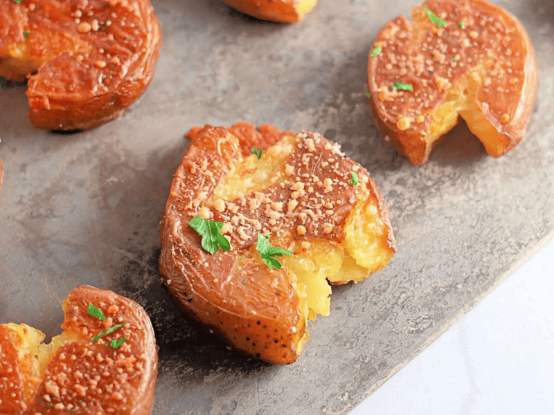 Several smashed, roasted baby potatoes garnished with herbs and grated cheese are arranged on a baking sheet.