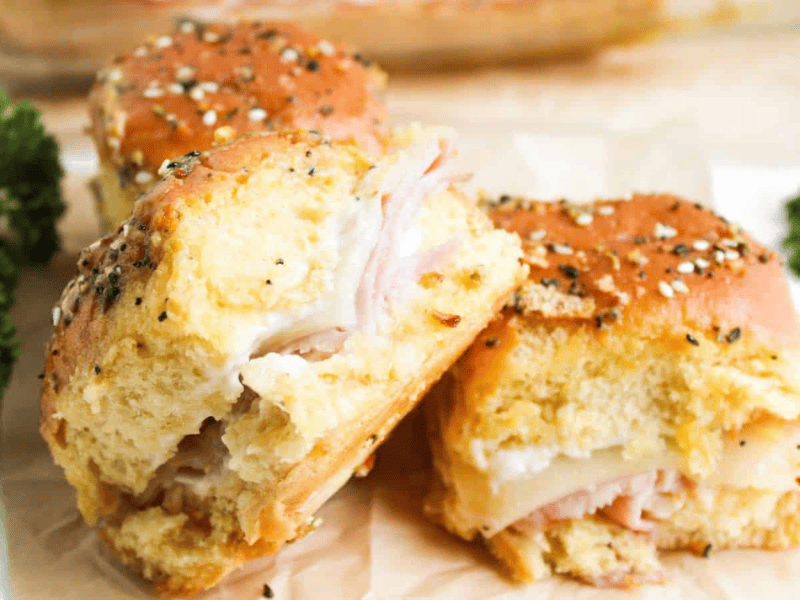 Three ham and cheese sliders on seeded buns are placed on parchment paper. Bright green parsley is visible in the background.