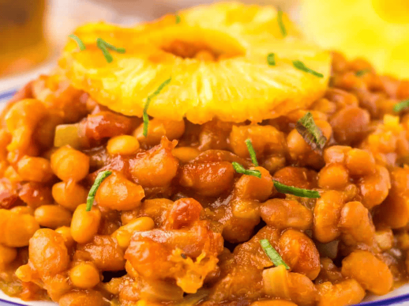 A plate of Hawaiian baked beans garnished with a pineapple slice and chopped herbs.