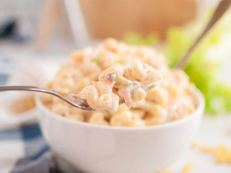 A close-up shot of a fork lifting a portion of macaroni salad, with a bowl of the salad in the blurred background.