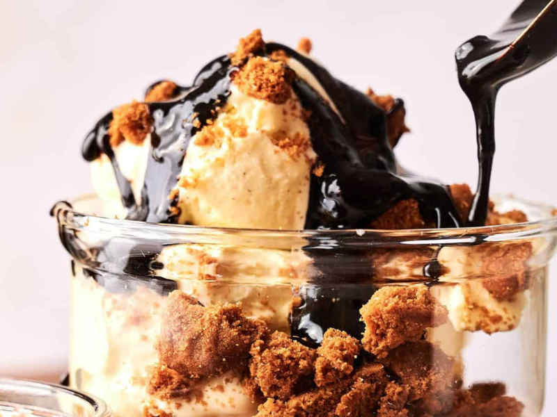 A glass filled with vanilla ice cream, crumbled cookies, and chocolate sauce being poured on top.