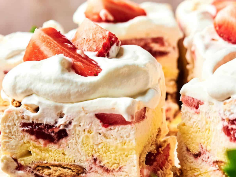Close-up of strawberry shortcake slices topped with whipped cream and fresh strawberries. Layers of cake, cream, and strawberries are visible.