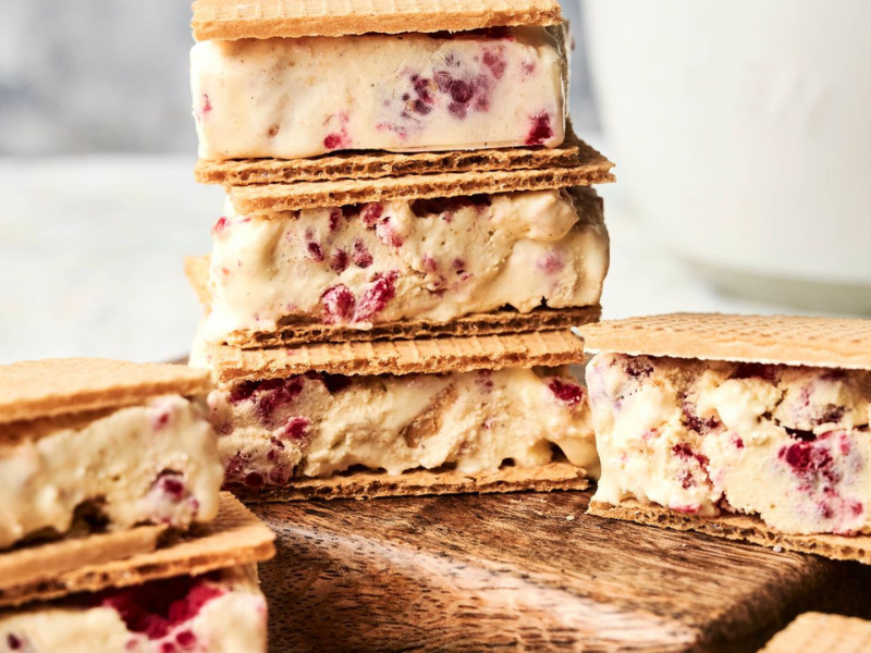 Ice cream sandwiches with wafer cookies and ice cream containing visible berry pieces, arranged on a wooden board with a white background.