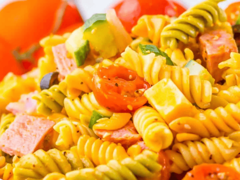 A vibrant pasta salad with fusilli, cherry tomatoes, bell peppers, black olives, and diced salami in a clear bowl.